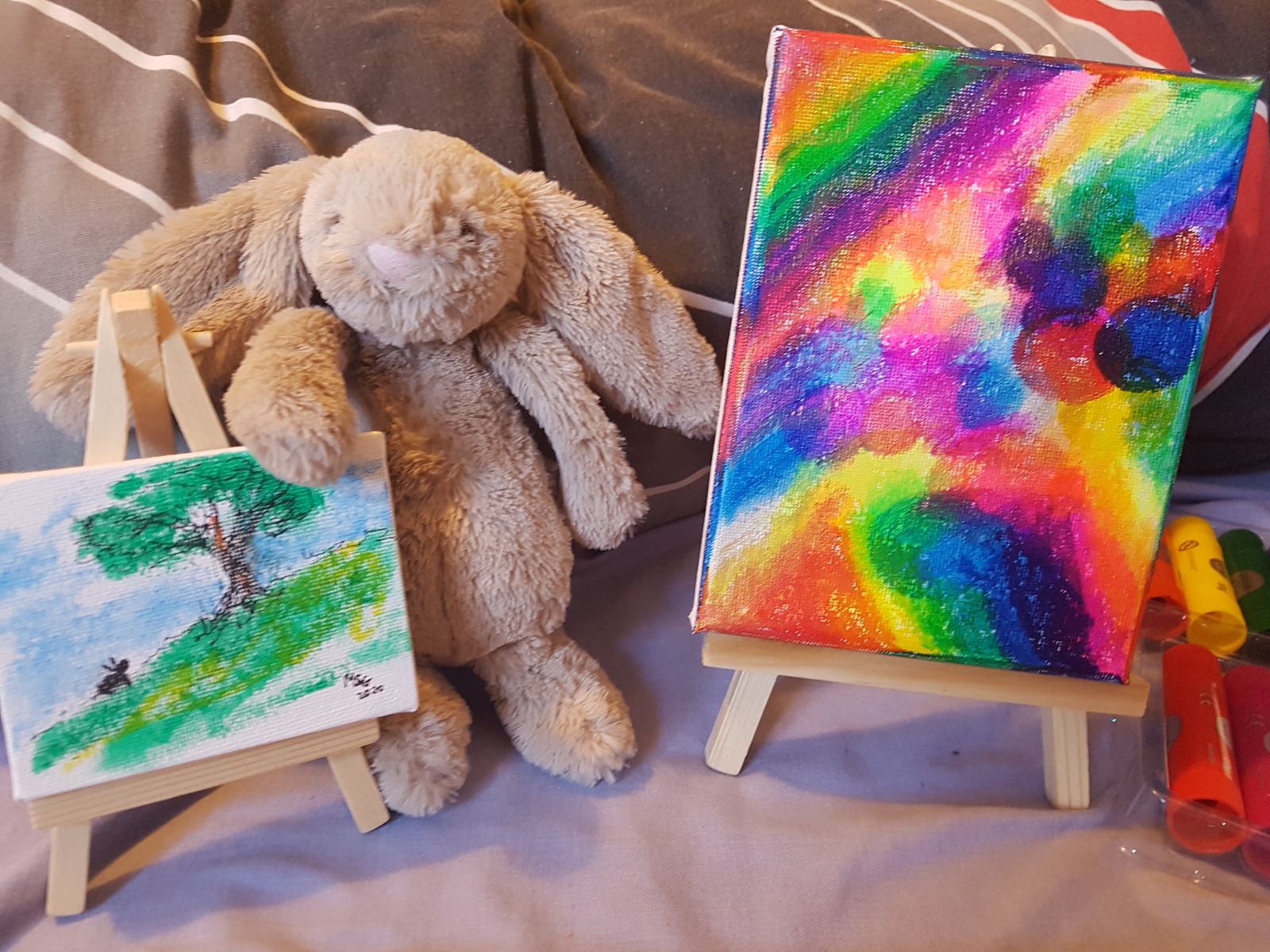 A toy rabbit is posed with another tiny rabbit painting placed on a tiny easel. A larger painting in rainbow colours stands on a slightly larger easel.