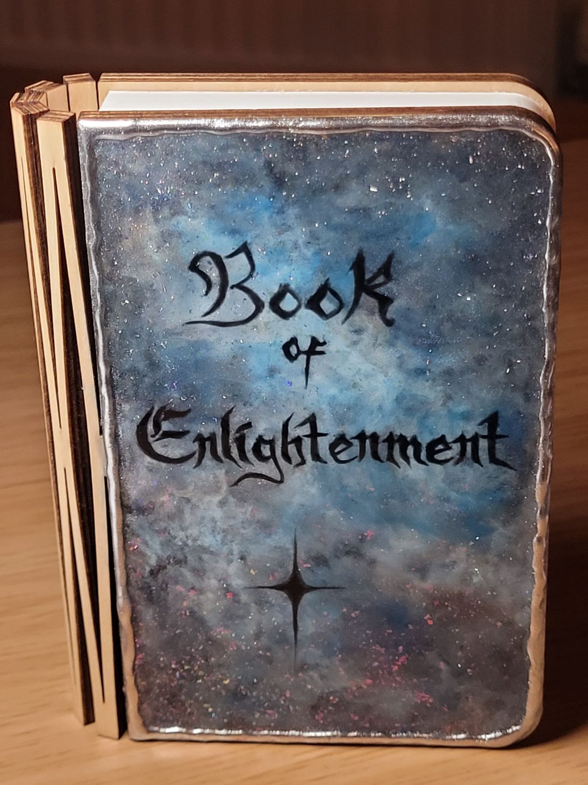 Book shaped lamp with a front cover that looks like blue and white clouds, with sparkling stars and silvered edges. The words 'Book of Enlightenment' are written across the centre in a gothic script.