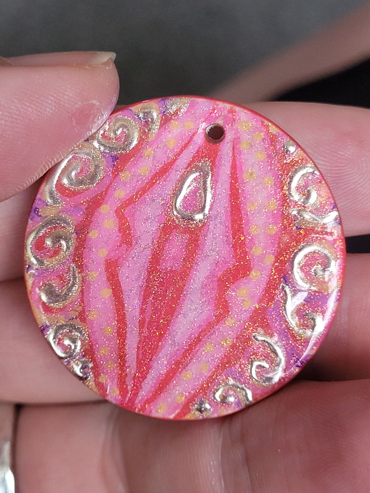 A hand painted badge in the approximate colours of the lesbian flag (reds, pinks, oranges and yellows), with an abstract design reminiscent of a vulva. The 'clitoris' and spirals of 'pubic hair' are gilded.