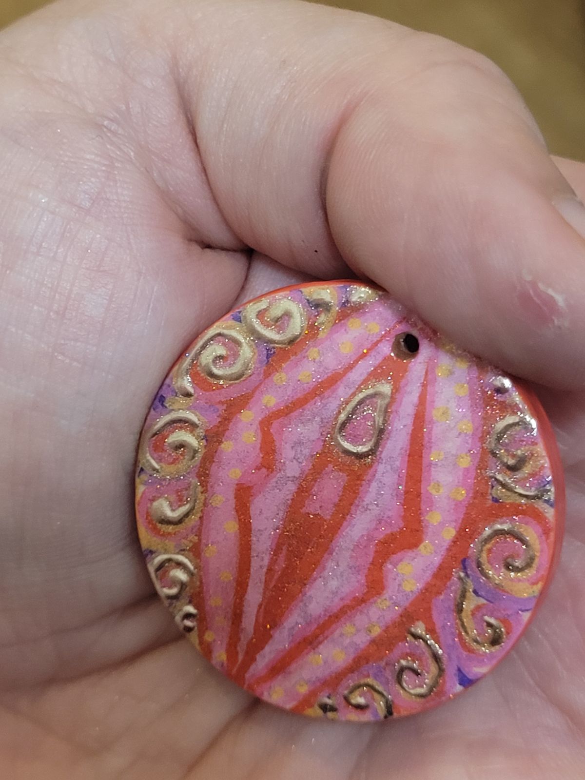 A hand painted badge in the approximate colours of the lesbian flag (reds, pinks, oranges and yellows), with an abstract design reminiscent of a vulva. The 'clitoris' and spirals of 'pubic hair' are gilded. Viewed from a slightly different angle so different colours show up.