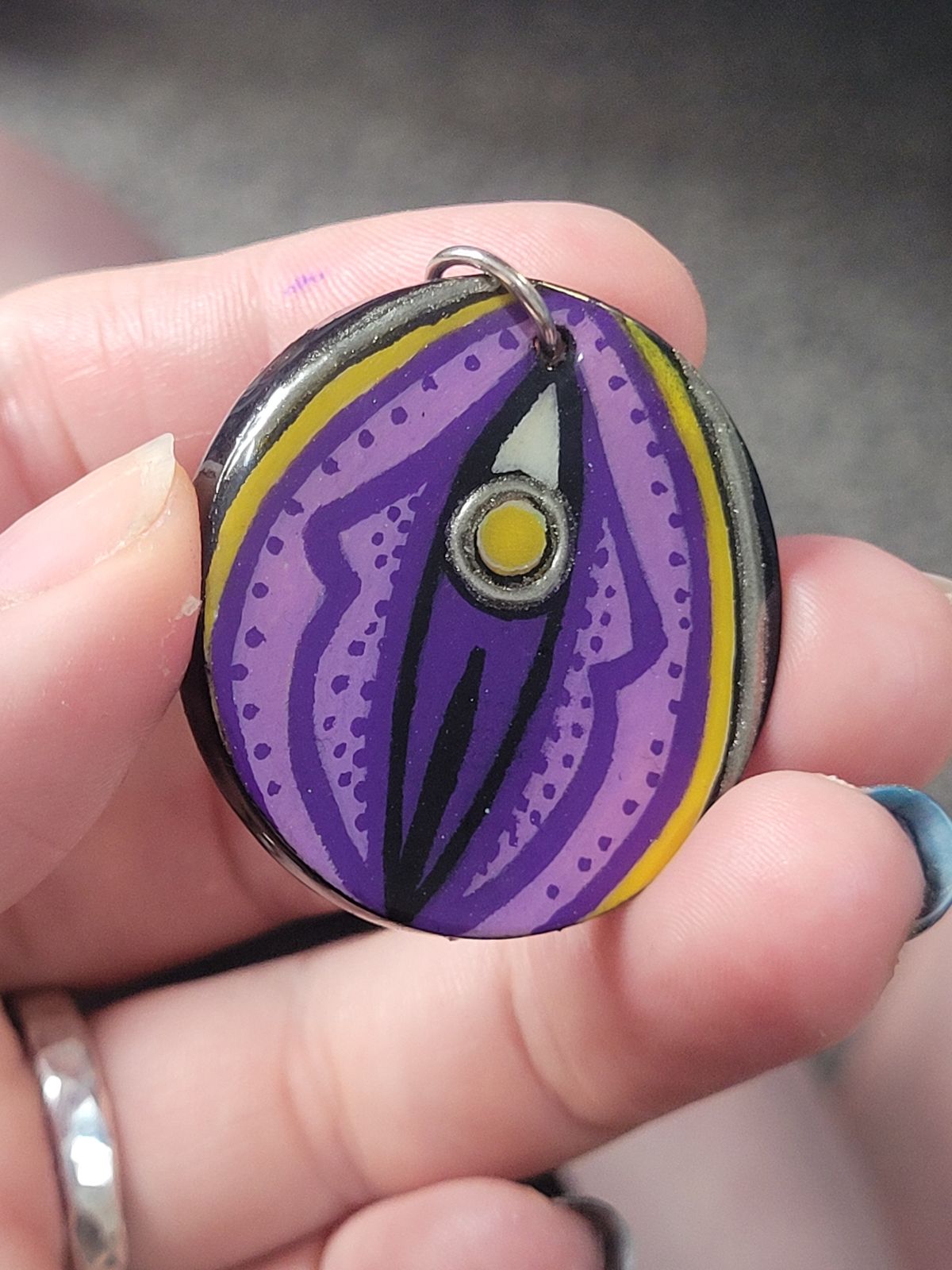 An abstract design reminiscent of a vulva, drawn in non-binary flag colours (black, white, yellow and purple). The 'clitoris' and a thin circle around the outer edge are gilded. A small steel ring has been added through a hole in the top.