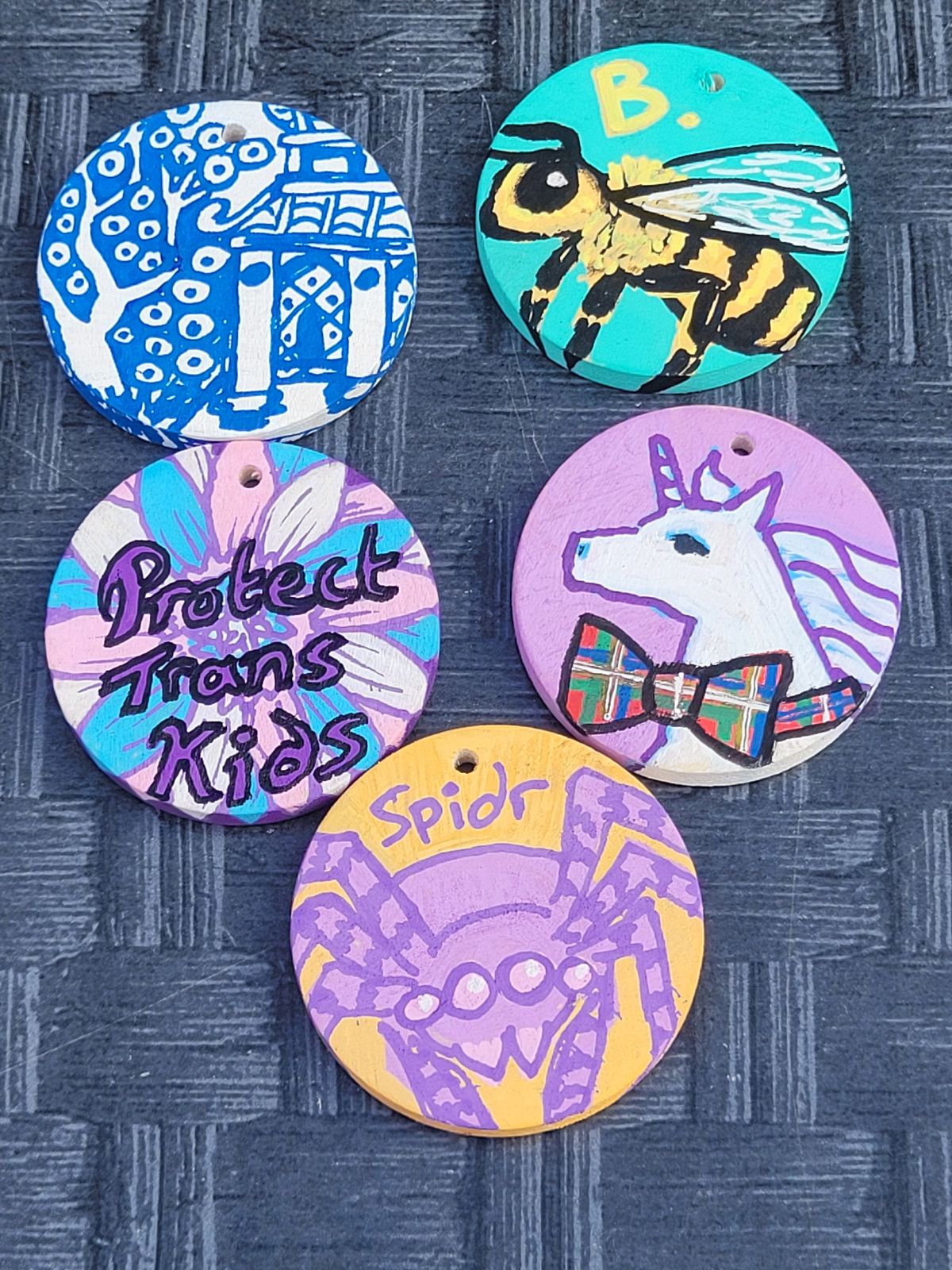 Badges depicting a unicorn with a plaid bow, a bee (labelled 'B'), a flower with 'protect trans kids', a blue and white china-inspired badge, and a lilac spider.