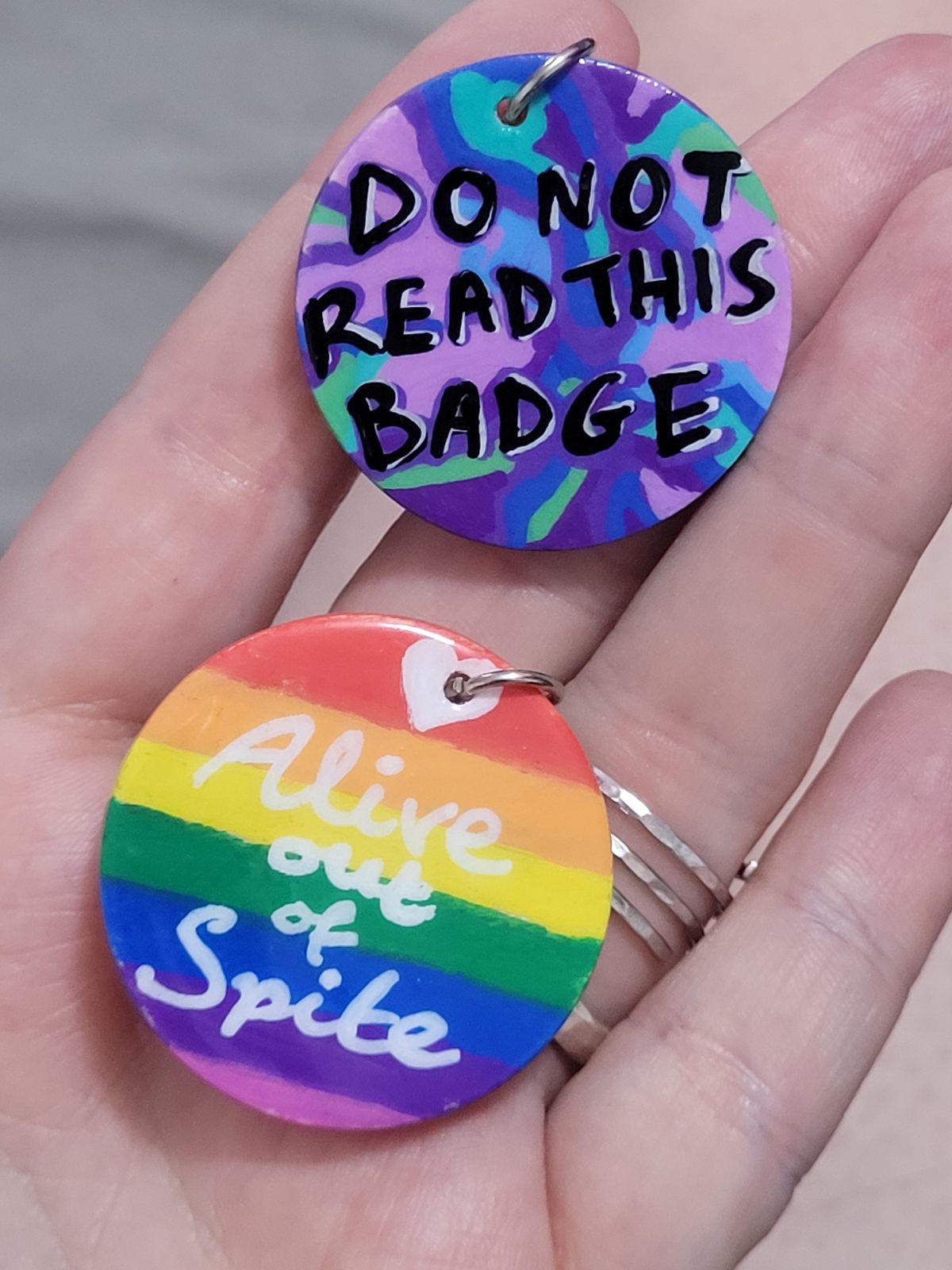 Text badges: 'Alive out of spite' on a rainbow background, and 'DO NOT READ THIS BADGE' on a colourful camouflage style background