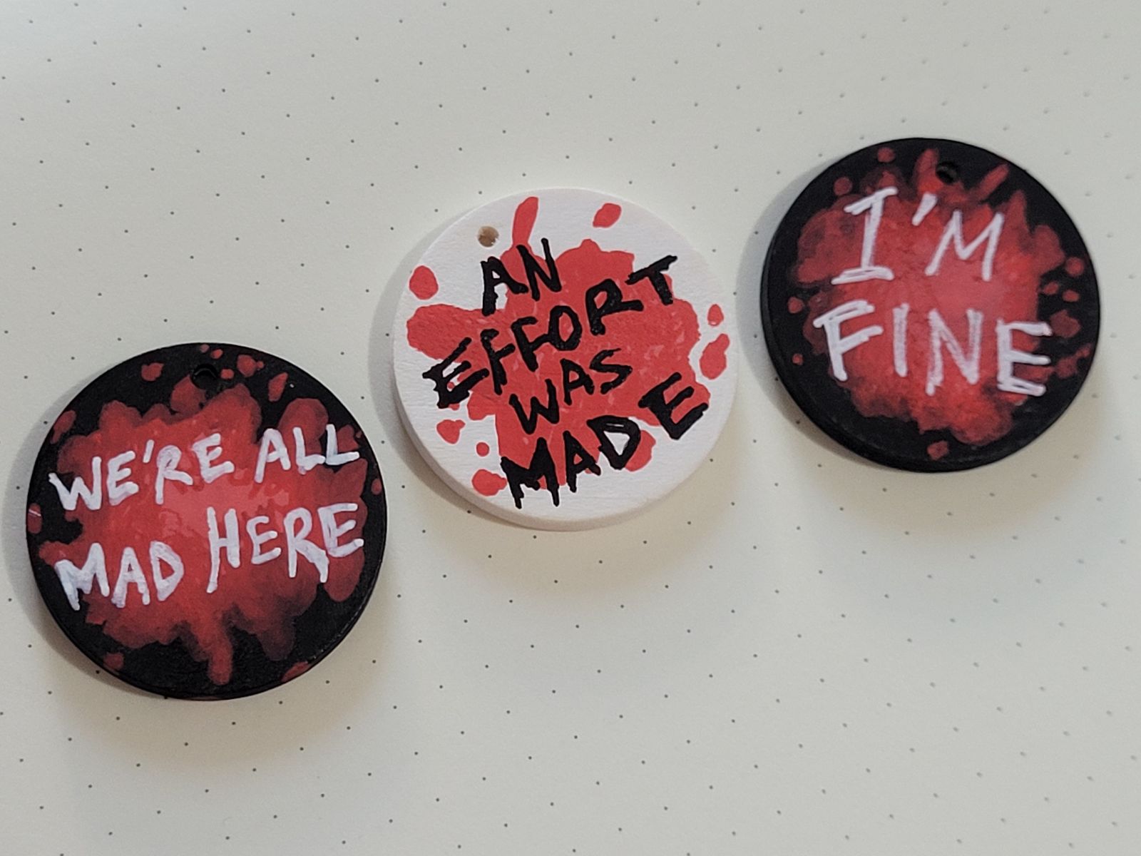 Three badges with a red 'blood splatter' background: 'WE'RE ALL MAD HERE', 'AN EFFORT WAS MADE', and 'I'M FINE'.