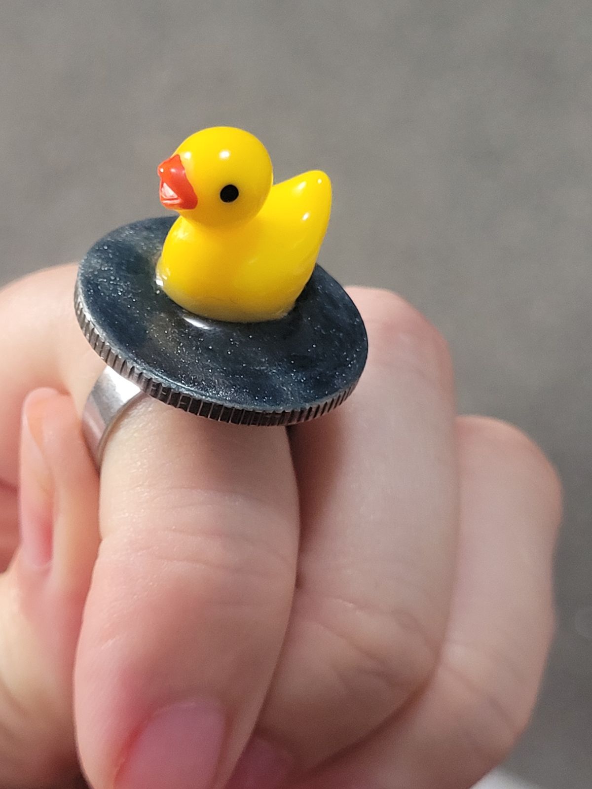 A small plastic duck sits on a circular base made to look like water, atop a finger ring.