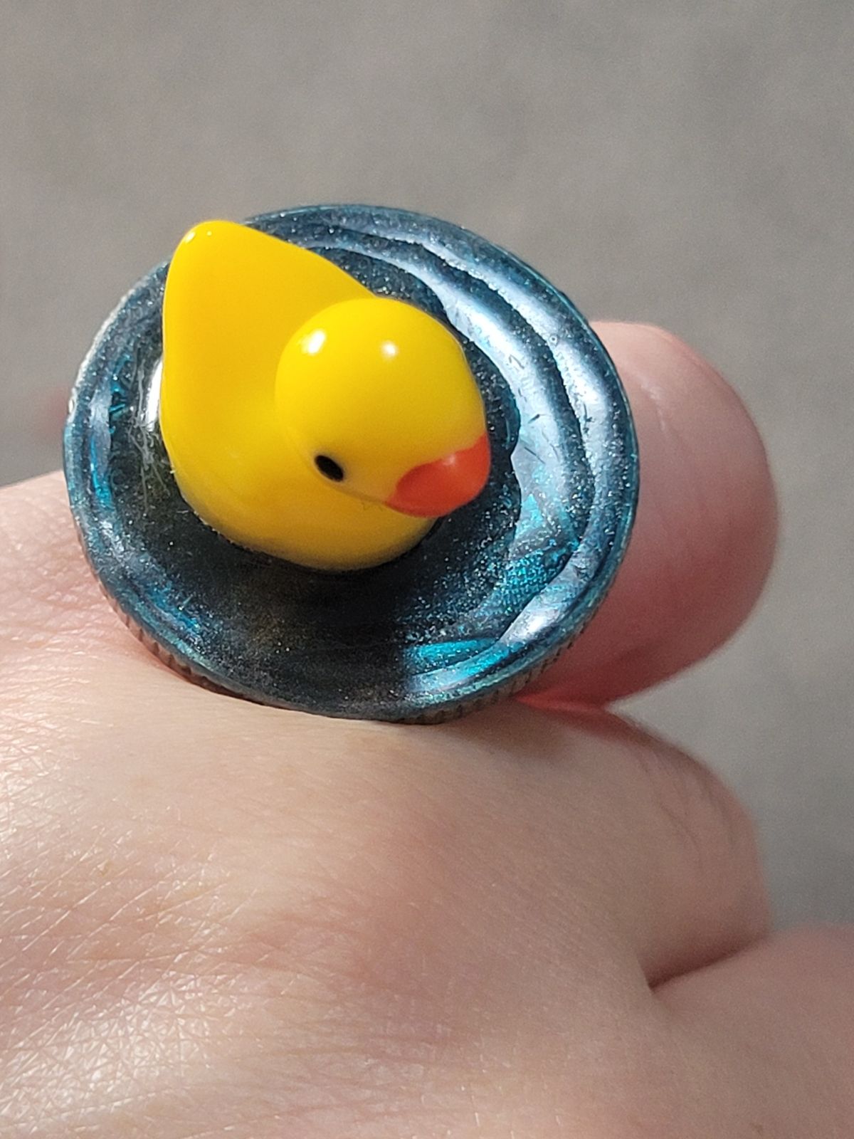 A small plastic duck sits on a circular base made to look like water, atop a finger ring.