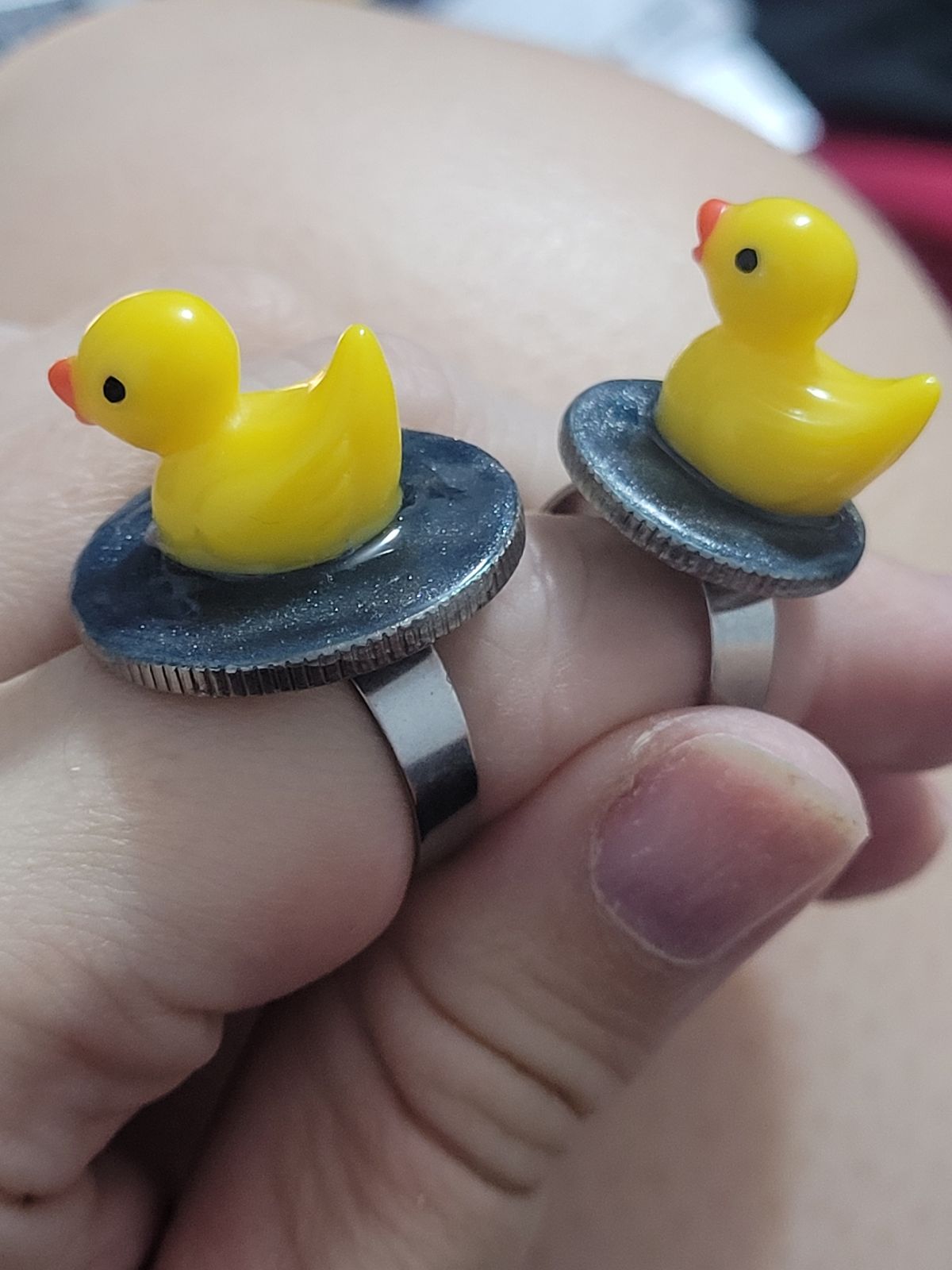 Two small plastic ducks sit on circular bases made to look like water, atop two finger rings.