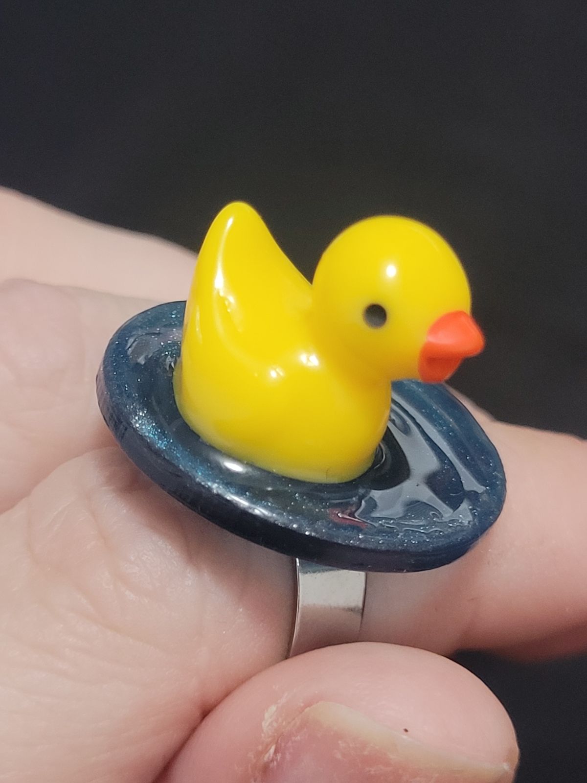 A small plastic duck sits on a circular base made to look like water, atop a finger ring.