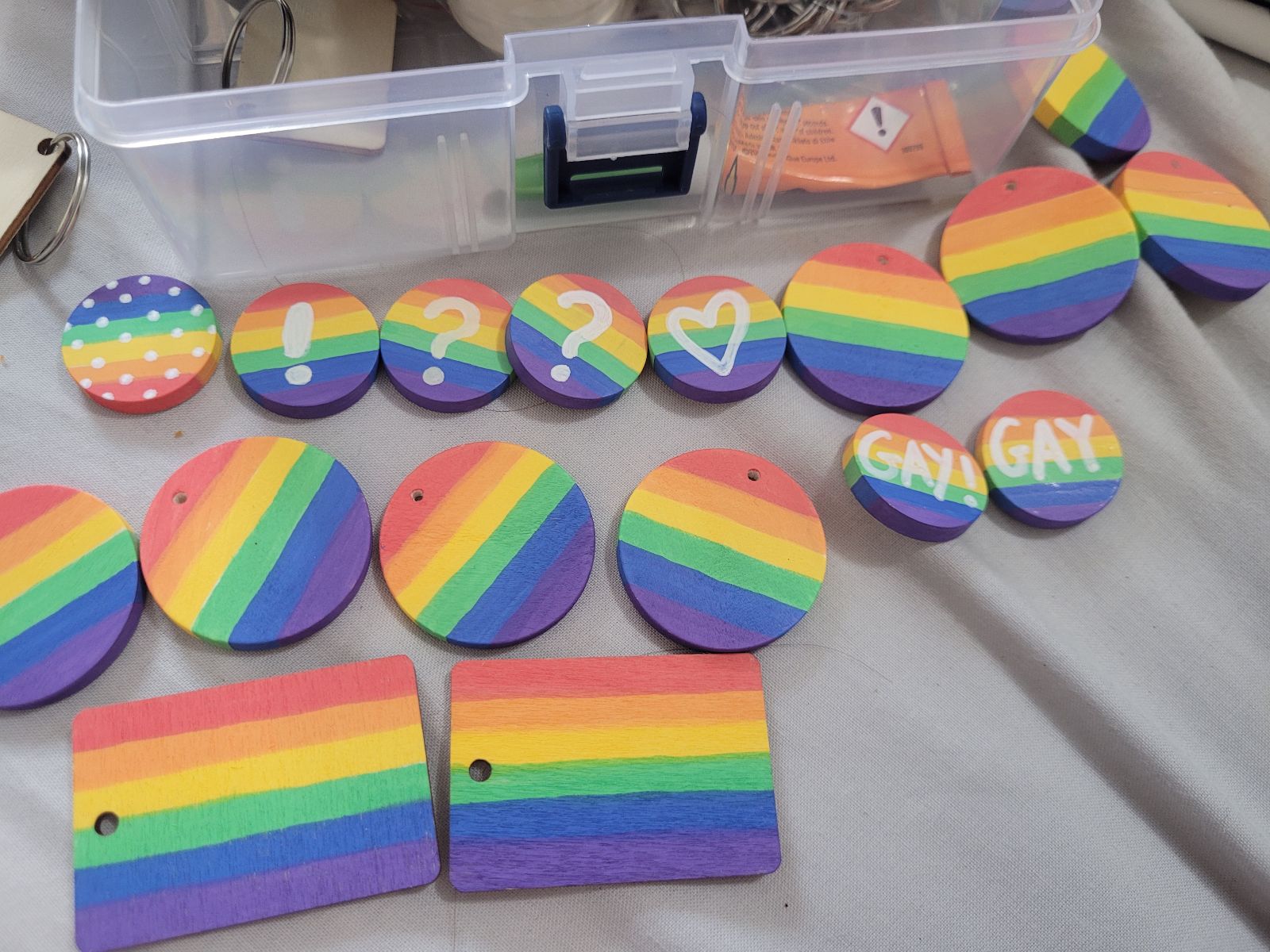 About a dozen rainbow badges in progress, laid out on a table. Some have white punctuation marks, or the word 'GAY' across them.