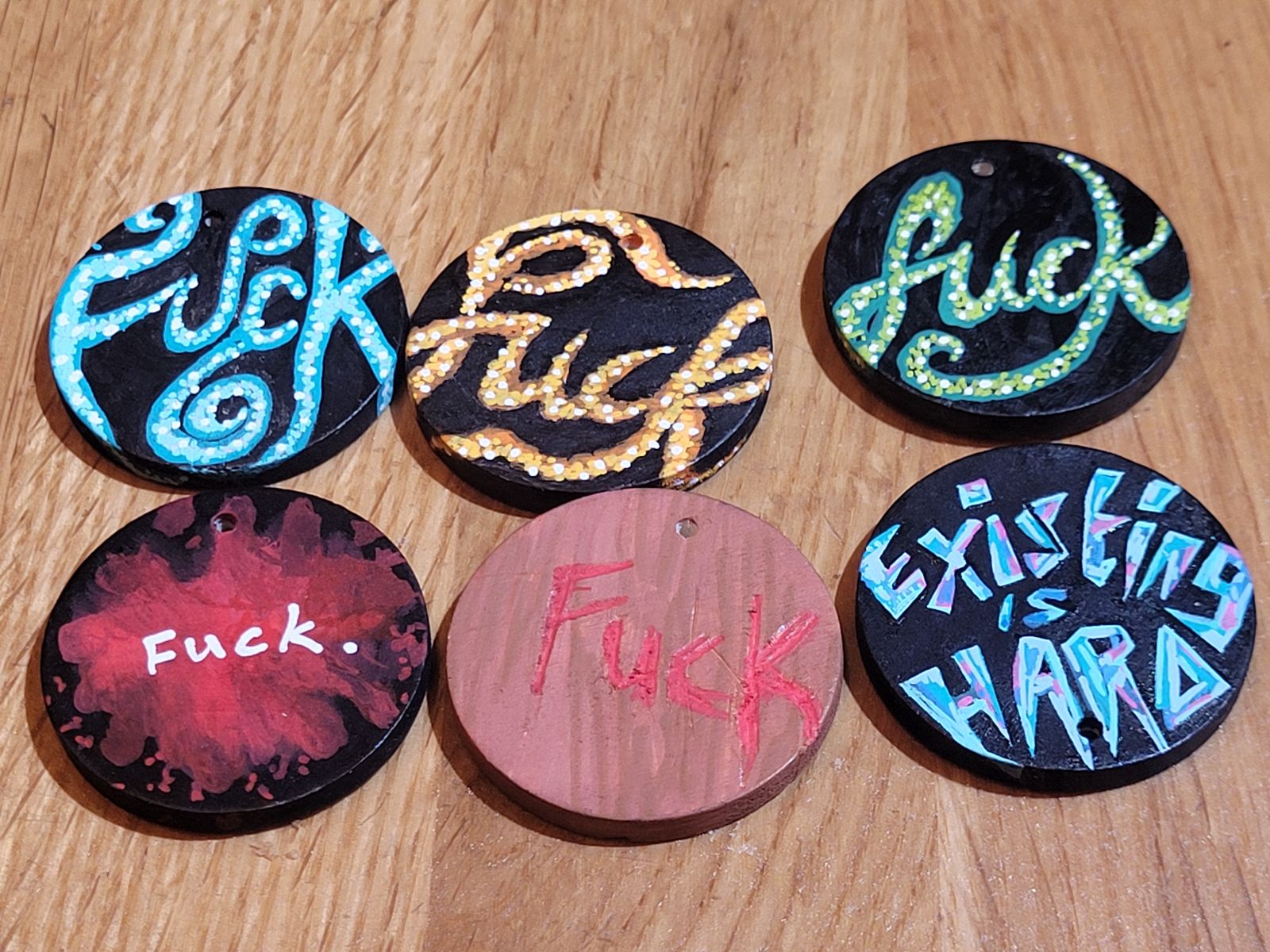 Three badges have text that looks like curling tentacles, reading 'Fuck' in various ways. Also one on a red blood-splatter background, one that looks like wood texture, and a last badge that reads 'Existing is Hard' in multicoloured jagged text.