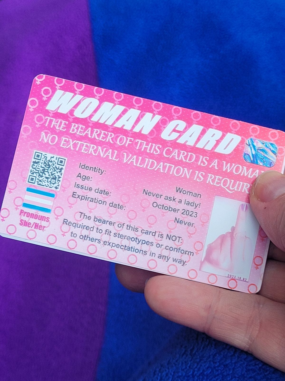 A mock ID card with a pink background reads 'Woman Card. The bearer of this card is a woman. No external validation required. There is an image of a hand with middle finger raised, wearing pink nail varnish. Smaller print reads 'The bearer of this card is not required to fit stereotypes or conform to others expectations in any way.'