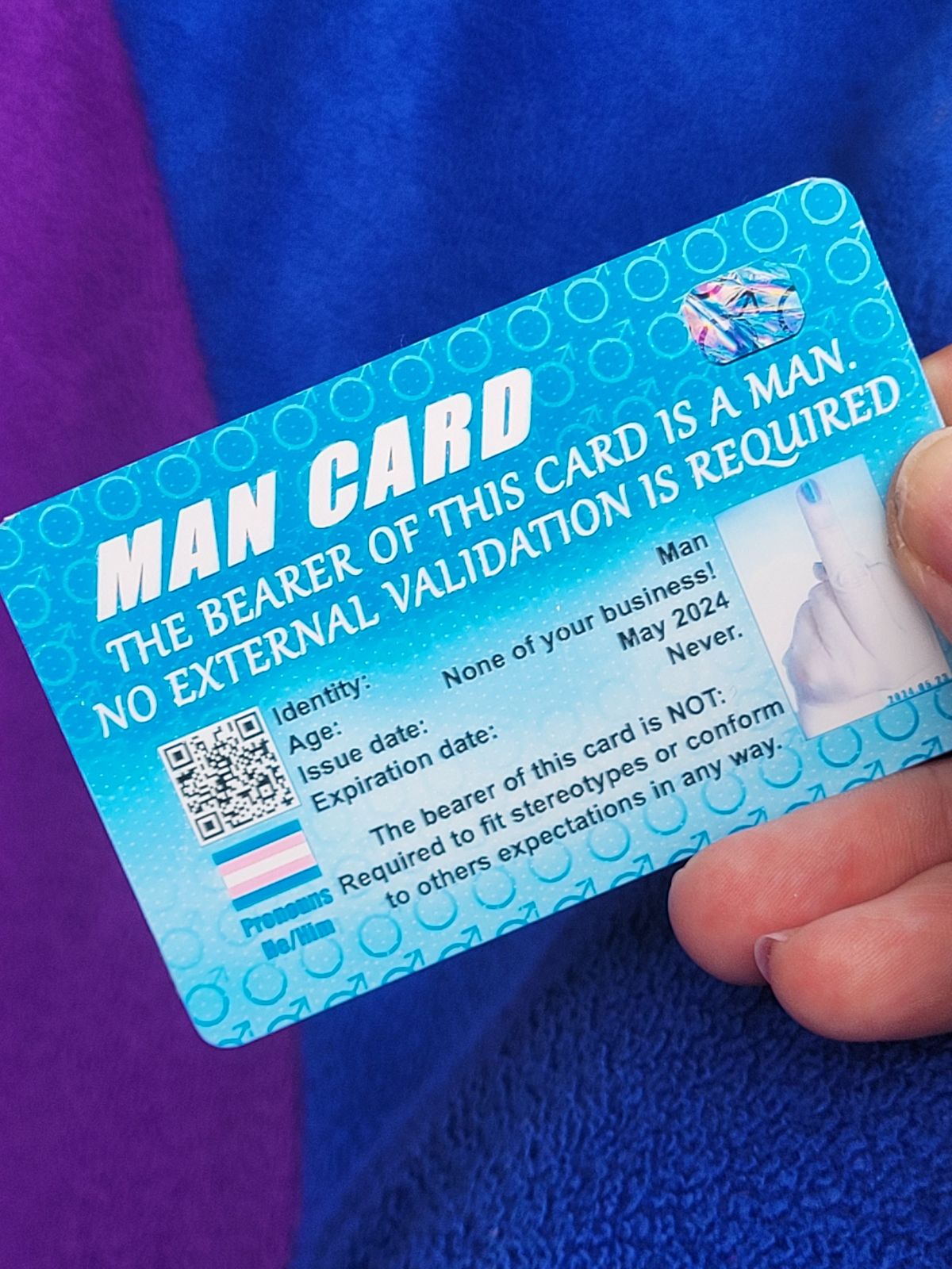 A mock ID card with a blue background reads 'Man Card. The bearer of this card is a man. No external validation required. There is an image of a hand with middle finger raised, wearing the same pink nail varnish as is seen on the woman card. Smaller print reads 'The bearer of this card is not required to fit stereotypes or conform to others expectations in any way.'